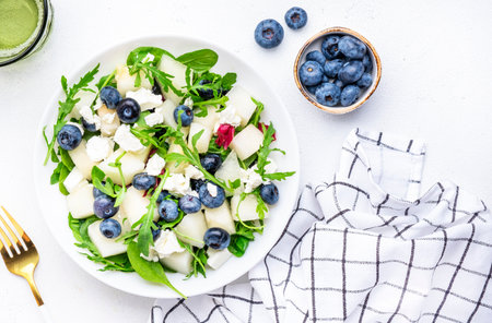 Delicious healthy salad with sweet melon, red onion, feta cheese, blueberries, arugula and mixed herbs, white table background, top viewの写真素材