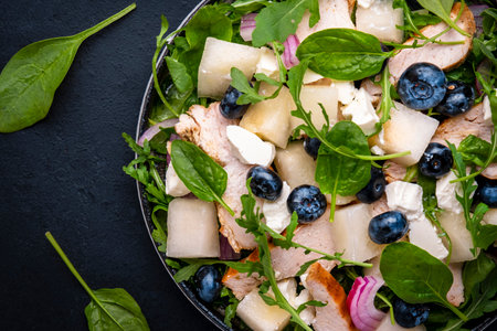 Gourmet salad with juicy sweet melon, grilled chicken, red onion, cheese, blueberries and mixed herbs, black table background, top viewの写真素材