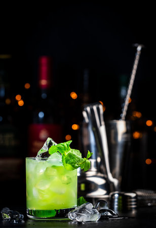 Green Negroni cocktail drink with dry gin, aperitif, gentian liqueur, fresh mint and ice. Black bar counter background, bar tools, bottlesの写真素材