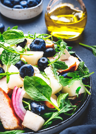 Fresh salad with sweet melon, grilled chicken breast, onion, feta cheese, blueberries and mixed herbs, black table background, top viewの写真素材