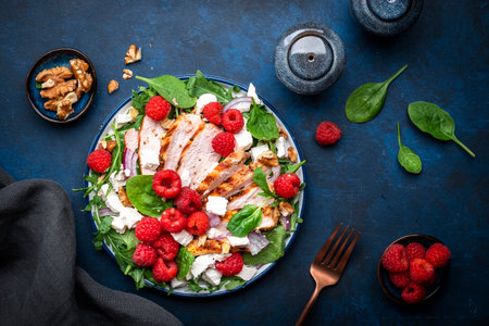 Tasty salad with raspberries, grilled chicken, cheese, onion, walnuts, spinach and arugula, blue table background, top viewの写真素材