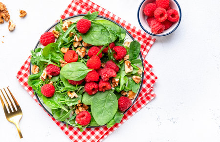 Tasty vegan salad with fresh raspberries, walnuts, spinach and mixed herbs, white table background,...