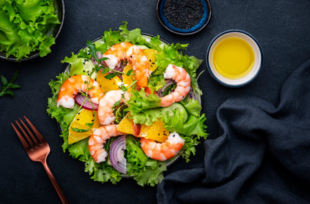 Fresh shrimp salad with orange, lettuce, cucumbers, red onion and sesame seeds with olive oil, black background, top viewの写真素材
