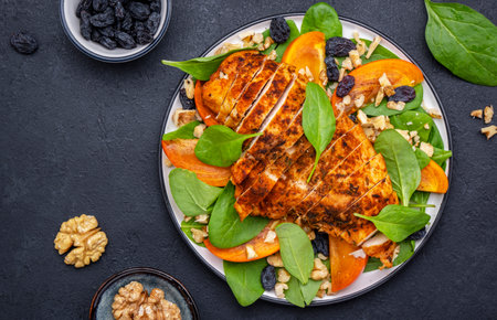 Winter persimmon salad with grilled chicken, walnuts, blue raisin and spinach, black background, top viewの写真素材