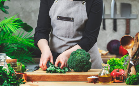 Chef picking broccoli leaves with his hands. Cozy kitchen with wooden table, kitchen utensils, vegetables, herbs and ingredients for cooking. Homemade food, cooking, recipes, food bloggingの写真素材