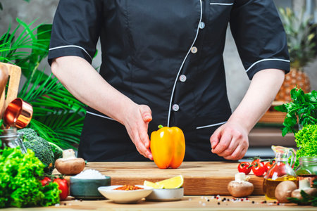 Chef in uniform holding orange paprika in hands.. Cozy kitchen with wooden table, kitchenware, vegetables, herbs and ingredients for cooking. Healthy vegan eating, culinary, recipes, food blogging comceptの写真素材