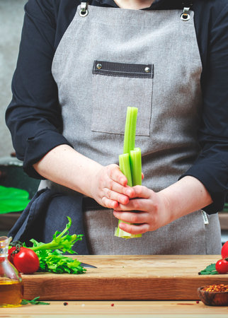 Home cook holding celery stalks in hands. Cozy kitchen with wooden table, kitchenware, vegetables,...
