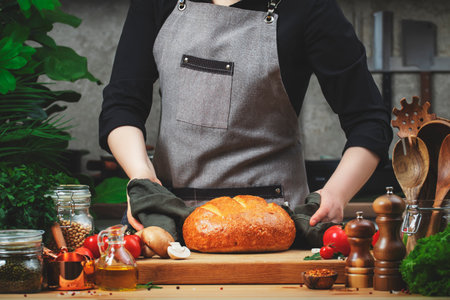 Chef holds freshly baked wheat bread in linen towel. Cozy kitchen with wooden table, kitchenware and cooking ingredients. Homemade baking, cooking, food bloggingの写真素材