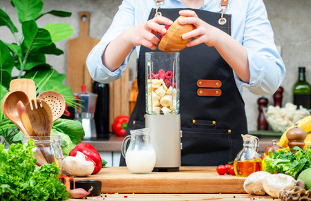 Chef pours frozen raspberries into blender with bananas to make milkshake. Cozy kitchen with wooden table, kitchenware, vegetables, herbs and ingredients for cooking. Healthy vegan eating, culinary, recipes, bloggingの写真素材