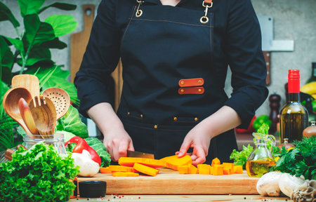 Cook cuts pumpkin pieces into cubes with knife. Cozy kitchen with wooden table, kitchenware, vegetables, herbs and ingredients for cooking. Healthy vegan eating, culinary, recipes, food bloggingの写真素材