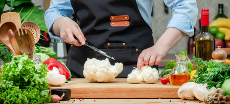 Cook cuts cauliflower into big florets. Cozy kitchen with wooden table, kitchenware, vegetables,...