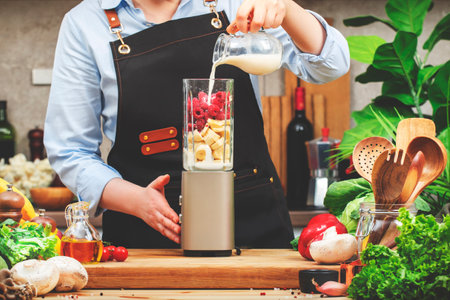 Cook pours vegan milk into blender with bananas and raspberry to make smoothie. Cozy kitchen with wooden table, kitchenware, vegetables, herbs and ingredients for cooking. Healthy vegan eating, drinkig, recipes, bloggingの写真素材