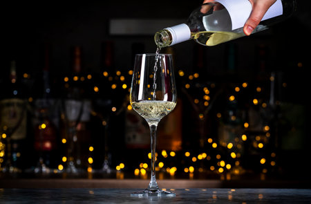White wine pours into glass and wine bottles on black bar background with lightsの写真素材