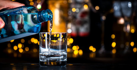 Dry gin pours in glass, strong drinks bottles on black bar counter background with lightsの写真素材