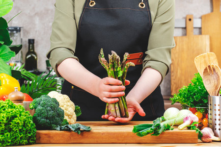 Chef cooking food in the kitchen. Cook holds fresh organic asparagus in hands. Wooden table, kitchenware, vegetables and herbs. Healthy vegan eating, recipes, blogging sceneの写真素材