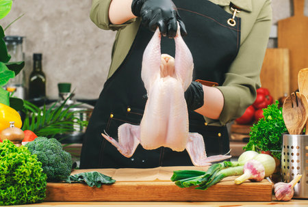 Chef cooking food in the kitchen. Cook holds whole raw chicken. Wooden table, kitchenware, vegetables and herbs. Healthy eating, recipes, bloggingの写真素材