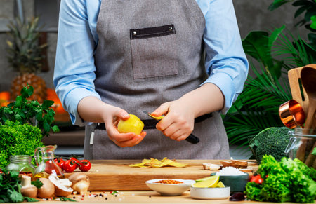 Chef peels potatoes for frying with vegetable peeler. Cozy kitchen with wooden table, kitchenware, vegetables, herbs and ingredients for cooking. Healthy vegan eating, culinary, recipes, food bloggingの写真素材