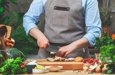 Cook preparing food in the kitchen. Chef cuts brown champignon mushrooms. Wooden table, kitchenware,...