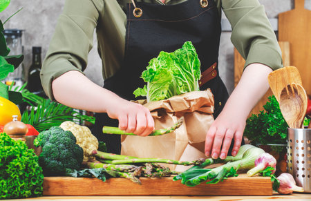 Chef cooking food in the kitchen. Cook gets green vegetables from paper bag. Wooden table, kitchenware, vegetables, herbs and ingredients. Healthy vegan eating, recipes, bloggingの写真素材