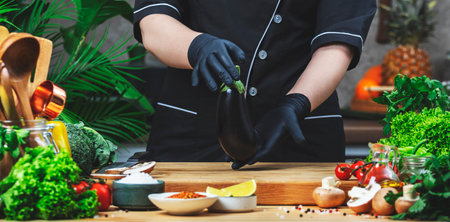 Cooking food in the kitchen. Chef holds fresh eggplant. Wooden table, kitchenware, vegetables, herbs...