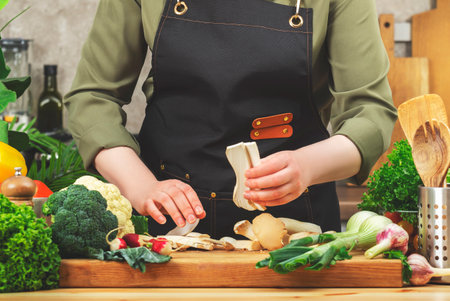 Cooking Food in the Kitchen. Cook holds chopped eringa mushrooms. Cozy kitchen with wooden table, kitchenware, vegetables, herbs and ingredients. Healthy vegan eating, culinary, recipes, bloggingの写真素材
