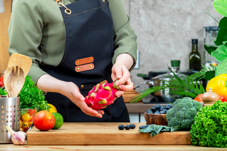 Cooking food in the kitchen. Chef holds pitahaya in hands. Wooden table, kitchenware, vegetables, herbs and ingredients. Healthy vegan eating, recipes, bloggingの写真素材