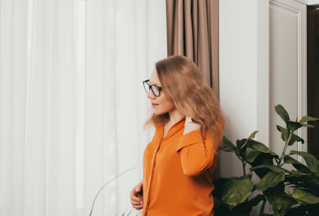 Joyful blonde woman wearing an orange shirt relaxes in her modern living room, enjoying a peaceful moment in her stylish apartment.の写真素材
