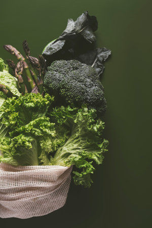 Freshly picked green vegetables and herbs are held in a reusable eco-friendly bag against a vibrant...