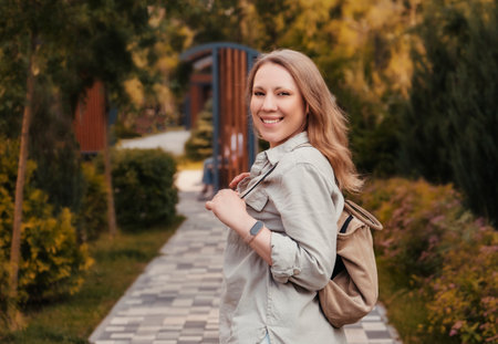 Blond woman with backpack takes a cheerful walk through a lush park adorned with greenery, showcasing a fitness bracelet on her wrist.の写真素材
