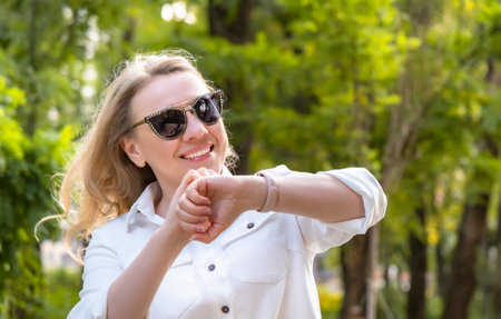 A happy woman takes a leisurely walk in a lush park, wearing stylish sunglasses and a fitness bracelet to monitor her fitness goalsの写真素材