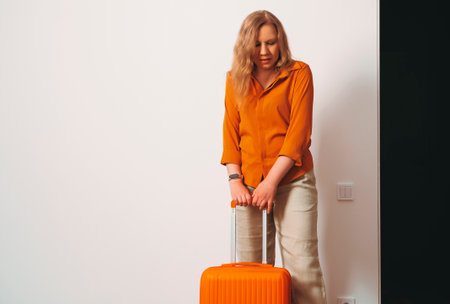 A woman wearing an orange shirt and beige pants stands beside an orange suitcase, appearing contemplative in a minimalist room.の写真素材