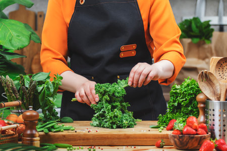 A woman prepares fresh leafy greens and other vegetables on a wooden counter in a bright, airy kitchen, healthy promoting vegan cooking.の写真素材