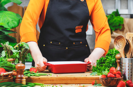 passionate woman chef holds a ceramic baking dish prepares a healthy meal in a cozy kitchen filled with fresh vegetables and farm herbs.の写真素材