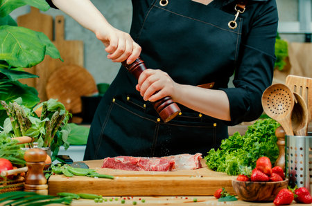 A woman chef sprinkle beef steak with spice from the mill prepares a nutritious meal using fresh farm herbs and vegetables in a cozy kitchen, highlighting her culinary skills.の写真素材