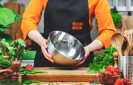 Chef woman in apron holds a metal bowl in hands prepares healthy dish using fresh vegetables in beautifully arranged kitchen filled with greenery.の写真素材