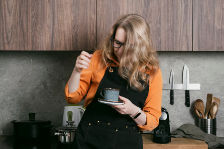 Blond woman prepares and enjoys a cup of espresso in her stylish kitchen, savoring a delightful coffee break moment.の写真素材