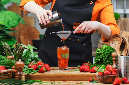 Chef woman skillfully pours a homemade basil strawberry mojito cocktail into a glass, surrounded by fresh greens and herbs on a wooden table.の写真素材