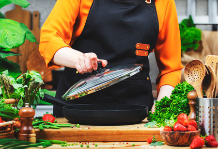 Chef woman in a black apron lifts a lid off a pan amid an array of fresh vegetables, prepping for a delightful vegan meal.の写真素材
