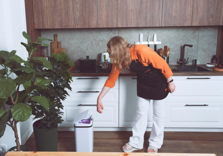 Woman in an orange shirt uses a smart sensor trash bin in a contemporary kitchen surrounded by plants and elegant furnishings.の写真素材