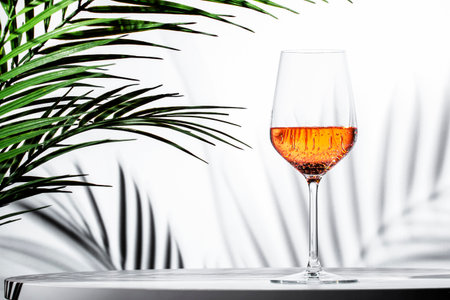 Elegant glass of rose pink wine served on a table with tropical palm leaves in the background for a refreshing experienceの写真素材