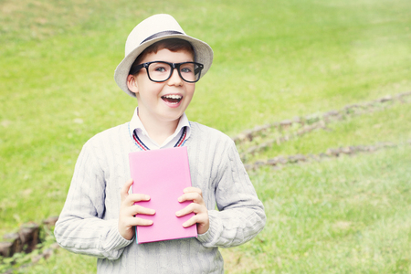 Portrait of young boy with eyeglasses, book and hat in the parkの写真素材