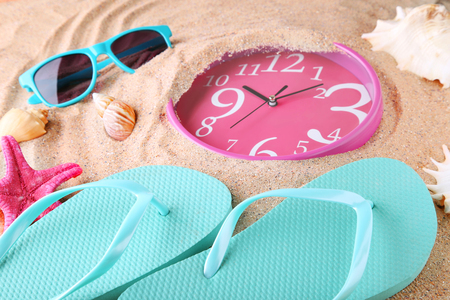 Pink clock immersed in sand with flips, sunglasses and seashells