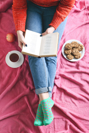 Woman with cup of coffee, book and plate of cookies sitting on pink plaidの写真素材