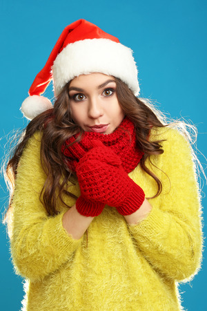 Beautiful young girl in yellow sweater, gloves, scarf and santa hat on blue backgroundの写真素材