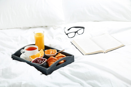Tasty breakfast in bed on wooden tray with book and glasses
