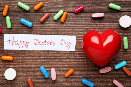 Inscription Happy Doctors Day with capsules and red heart on wooden tableの写真素材