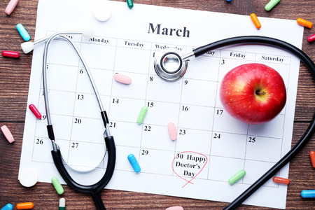 Inscription Happy Doctors Day with pills, stethoscope and red apple on march calendarの写真素材