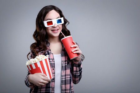 Young beautiful woman holding bucket with popcorn and paper cup on grey background