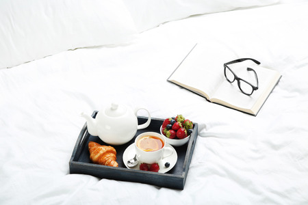 Tasty breakfast in bed on wooden tray with book and glassesの写真素材
