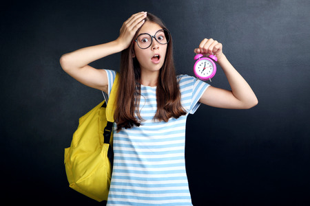 Young girl with backpack and alarm clock on blackboard background